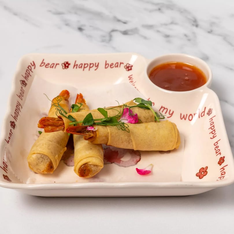 Crispy Thai shrimp spring rolls with sweet chili dipping sauce, garnished with pea shoots and flower petals.