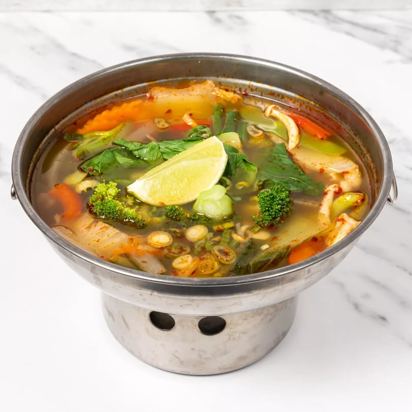 A metal pot of Jai Dee Restaurant's Thai soup with vegetables, mushrooms, and a lime wedge.