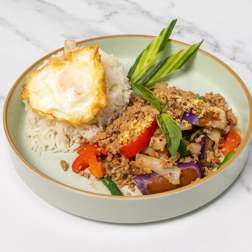 Thai dish with rice, fried egg, ground meat, eggplant, bell peppers, and basil on a light green plate.