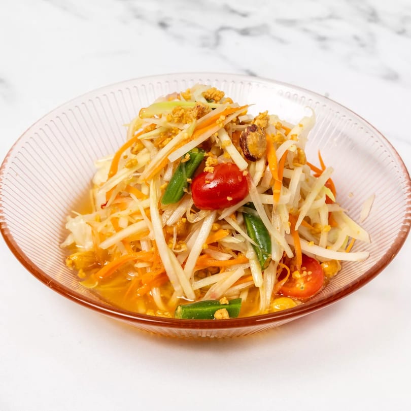 Som Tum, a Thai green papaya salad with tomatoes, green beans, and peanuts in a clear bowl.