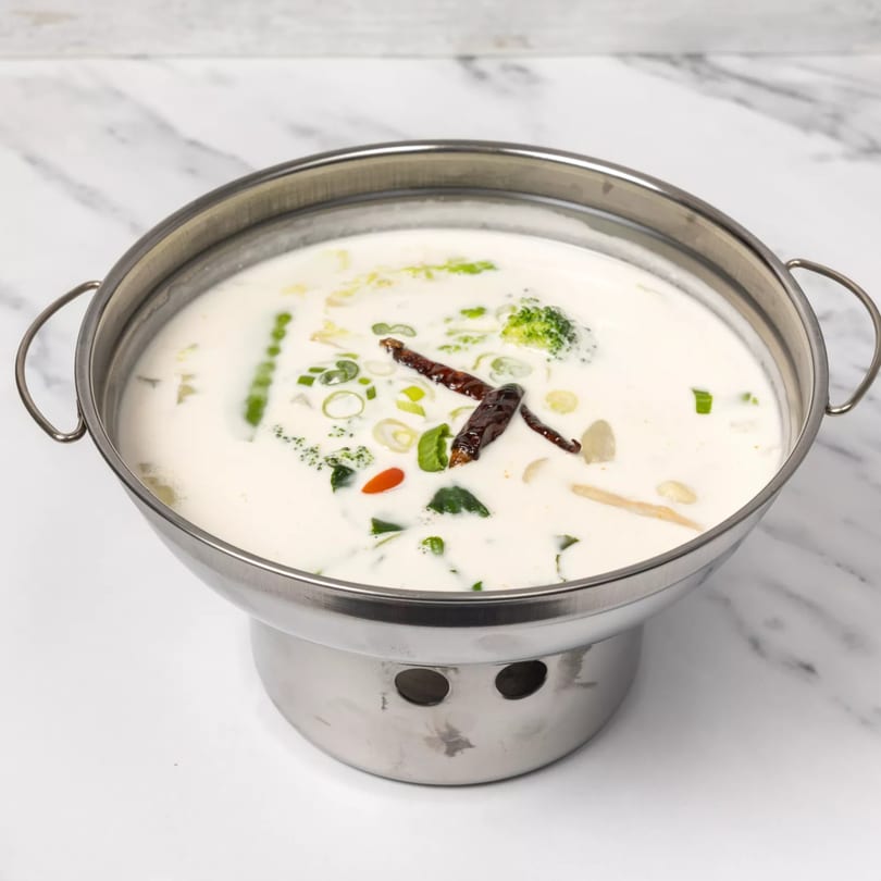 A stainless steel pot of creamy Thai soup with broccoli, green onions, and dried red chilies.